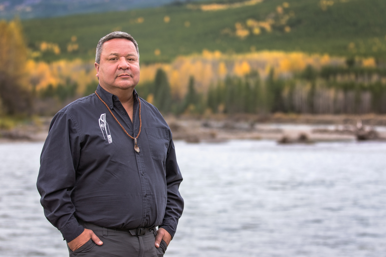 Kirby Muldoe, an indigenous man with short grey hair in a navy shirt, standing in front of a river. Photo by Carla Lewis Photography
